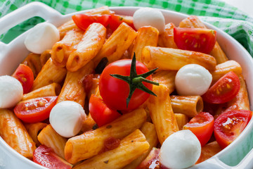 Penne pasta with sauce, mozzarella cheese and cherry tomatoes in casserole on white table.