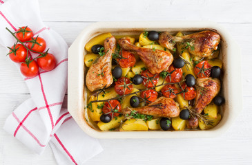 Baked chicken drumsticks in red dish. Cooked with cherry tomatoes, black olives, rosemary and potatoes. White wooden table, top view.