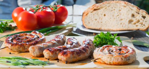 Assorted grilled sausages served on wooden plate over wooden table with vegetables and bread on background. Selective focus.