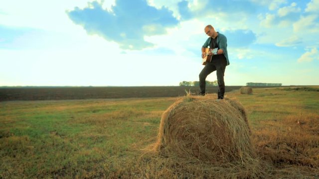 Thoughtful handsome guy playing guitar, sitting on a haystack. The concept of loneliness, happiness and a gay cowboy. Russian field and nature