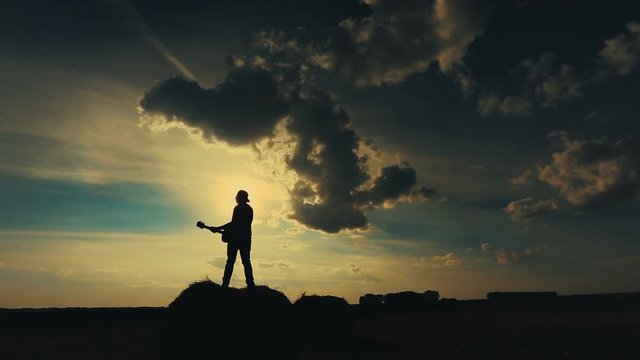 Thoughtful handsome guy playing guitar, sitting on a haystack. The concept of loneliness, happiness and a gay cowboy. Russian field and nature