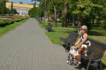 Fototapeta premium Woman resting on bench in the city park of Vrnjacka Banja in Serbia, while her dog is playing