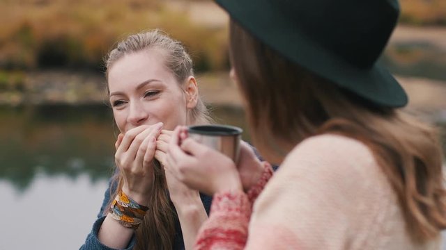 One girl plays harmonica and another girl listens music and drinks hot tea in the autumn forest near the river