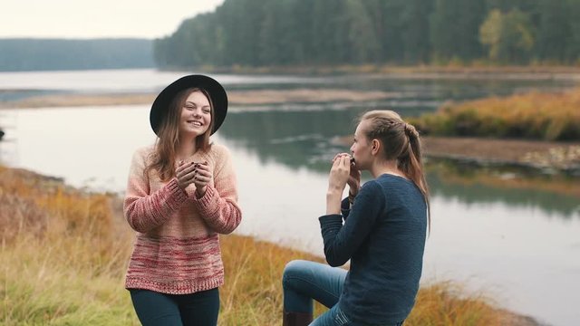 One girl plays harmonica and another girl listens music and drinks hot tea in the autumn forest near the river