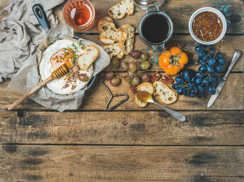 Camembert cheese in small pan, honey, fig jam, persimmon, grapes, pecan nuts, grilled baguette slices and glass of rose wine over rustic wooden background, top view, copy space