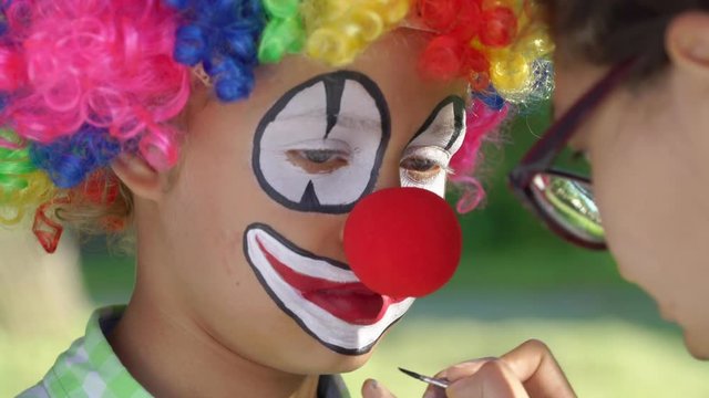 boy wearing funny clown and professional artist makes a children's make-up outdoors