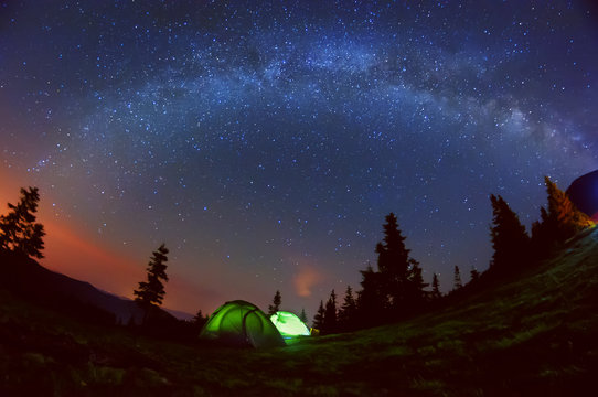 Night photo of the sky and the Milky Way. Wildlife, in a clearing in the mountains there are two glowing tent among the tall silhouettes of dark firs
