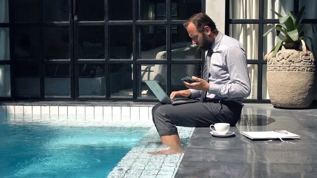 Businessman working with smartphone and laptop by pool in outdoor villa
