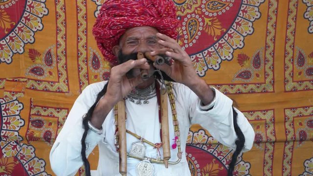 MS Hand-held Rajasthani elderly male starts to play the flute and then salutes in front of a colourful fabric tent