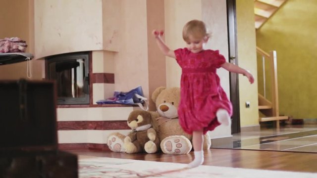 Adorable blonde cutie of 3 years old in an elegant red dress. Little girl comes to the living room with the lollipop, than throws it away, comes to the chest with toys closes it, and runs away.