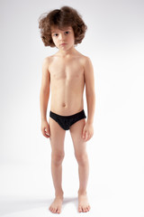 Naked curly-haired boy in shorts standing on the floor and looking into the camera. Gray background.