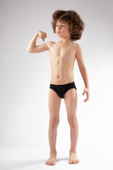 Curly boy in shorts showing muscles, bending the arm at the elbow. He looks into the camera. White background.