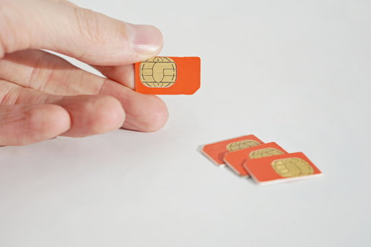 Isolated Photo Of Male Hand Holding Red SIM Card Above A Group Of 3 Other SIM Cards Used In The Mobile Phones (cell Phone Devices) With A Focus On Their Golden Micro Chip 