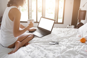 Young lady freelancer sitting on the bed in hotel room in front of window and working in laptop. Wide angle.