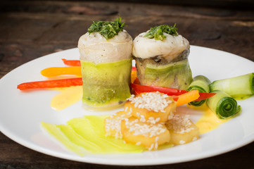 Fish rolls wrapped in zucchini with roasted potatoes sprinkled with sesame seeds and slices of bell pepper and cucumber. Wooden rustic background. Top view. Close-up