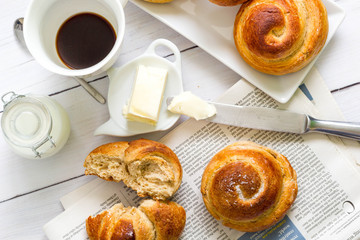 Brioche with butter, milk, a cup of coffee on newspaper. White table, top view 