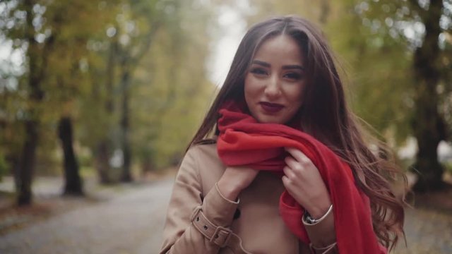 Attractive brunette girl with wonderful hairstyle and makeup, in stylish autumn look walking in the park, turns to camera and gives a charming smile. Park surroundings, trees, fallen leaves, benches.