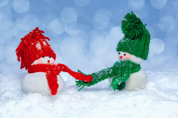 Two snowmen wearing a knitted hats  and scarves on snowy
 blurred background hold hands.   Winter holiday decor