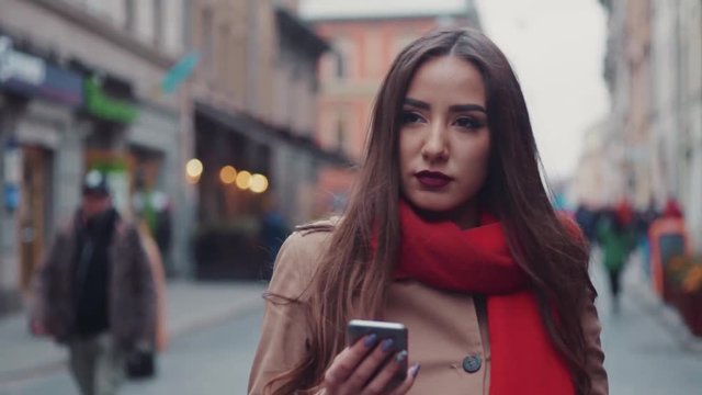 Gorgeous young lady with beautiful lips, long hair, bright red scarf and passionate glance. Attractive European woman walking through the crowd of ordinary people and using her phone.