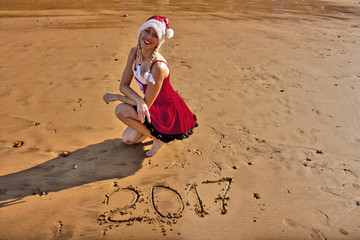 Woman in red dress and Christmas hat on the beach with drawing on the sand digits 2017