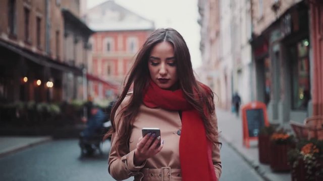 Close up view of attractive stylish girl uses phone while wondering in the city. Autumn wind playing with her long beautiful brunette hair and vivid red scarf. Forever young. Business lifestyle.