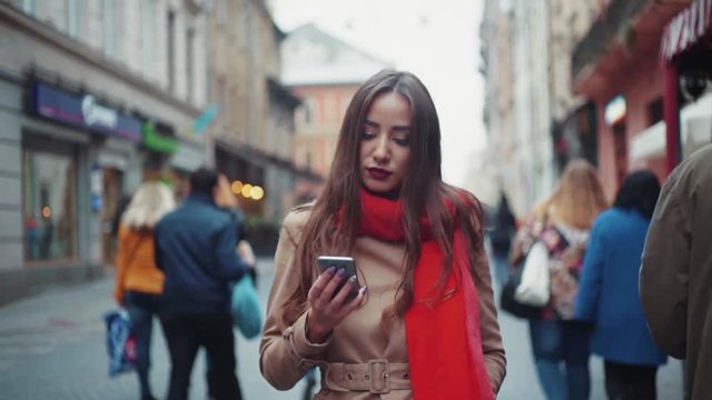 Beautiful young girl with red lips, in a warm autumn coat and vivid red scarf in the urban surroundings. Attractive brunette girl walking in the city center, and typing the message on her phone.