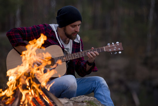 young handsome attractive bearded model man plaing guitar in forest with bonfire. Casual man, life style. Beautiful view on forest and rock - Powered by Adobe