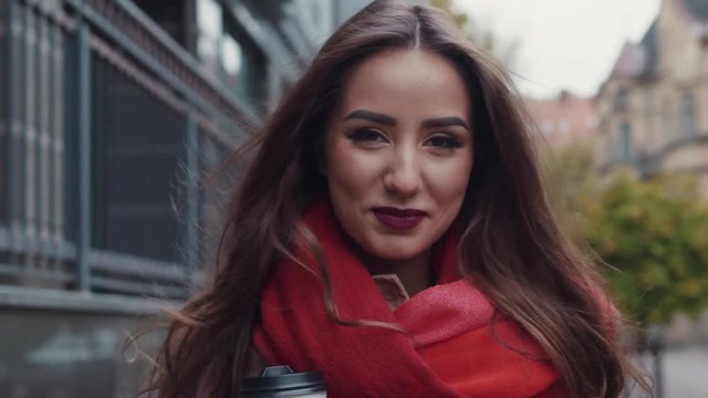 Attractive smiling girl in a vivid red scarf, with long hair and perfect makeup. Amazing young woman happily wonders in the autumn city. Freedom. Urban style. Stylish look. Happy moments.