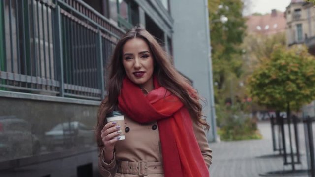 Beautiful young girl with an attractive appearance, adorable look, curly hairstyle, scarf and autumn coat in the city. Amazing view of business lady with a cup of coffee walking down the city street.