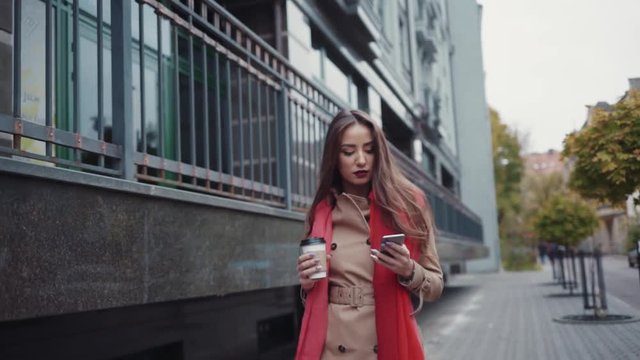 Attractive young girl in an autumn coat and bright red scarf. Amazing business lady having coffee and chatting on her phone, while walking down the city street.