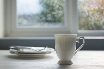 White cup and empty plates on white wooden table, with blurred window on the background, with beautiful bokeh