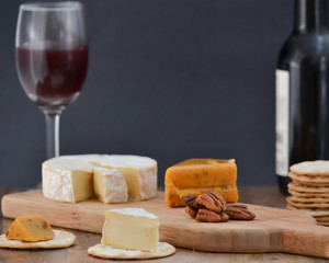 Brie, pepper jack, pecan halves and crackers on wooden cheese board, with one bottle and one glass of red wine, against dark background - celebrating alone concept