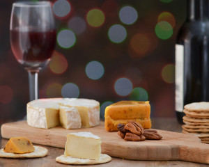 Brie, pepper jack, pecan halves and crackers on wooden cheese board, with one bottle and one glass of red wine, against dark holiday lights background - celebrating alone concept
