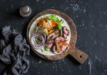 Tortilla with beef steak, marinated carrots and cabbage. Delicious lunch snack. On a dark background, top view