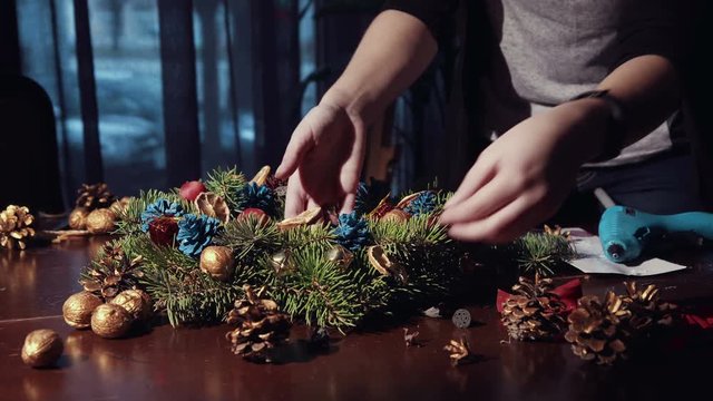 Crop close up hands decorating Christmas wreath with pine cones and ribbons using hot glue gun.