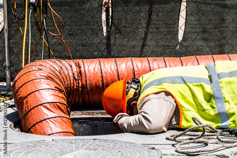 Wall mural worker wearing safety gear and looking down manhole in an urban setting.