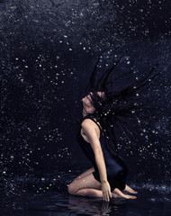 Fototapeta premium Pretty brunette woman in black dress under the water drops in aquastudio sitting in water on the knees