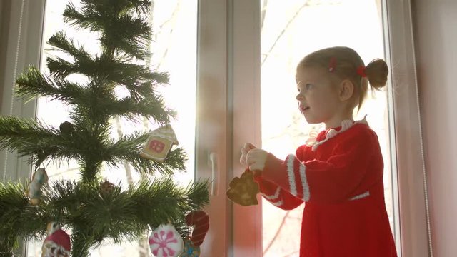 Little girl in a red dress adorning Christmas tree with gingerbread in the shape of Christmas angel against the bright window, standing on the windowsill.
