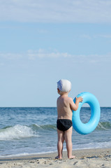 Kid in swimming trunks and a cap with a swimming circle stands on the seashore with his back