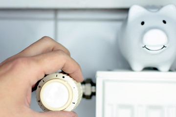 Close-up Of a Mans Hand Adjusting Thermostat With Piggy Bank On Radiator