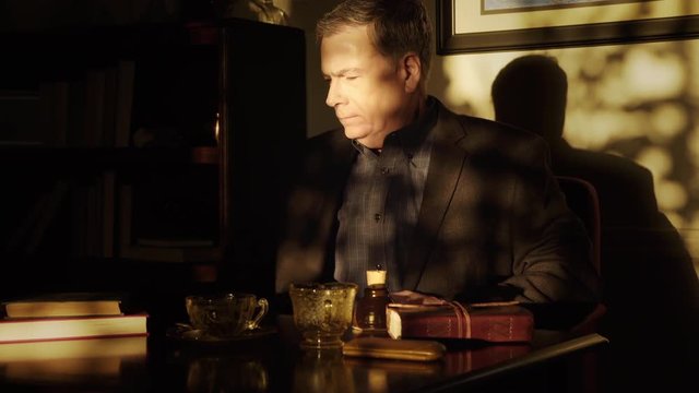 An mature man enters his office, which is lit only by early morning sunbeams, sits and starts to look over some paperwork.
