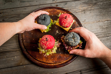 Small American colored burgers on a rustic wooden table, a snack at one bite. Man and woman take two burgers from board, hands in the picture, top view