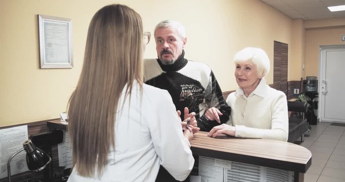 Elderly patients talking with nurse receptionist at hospital reception desk 4k video. Couple visits clinic or diagnostics center 