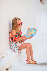 Little girl with map of island outdoors in old streets an Mykonos. Kid at street of typical greek traditional village with white walls and colorful doors on Mykonos Island, in Greece