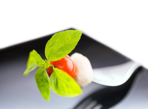 Italian salad. Cheese mozzarella, basil and tomatoes on a fork on a black plate. In small DOF.