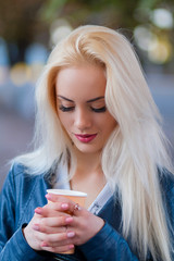 Beautiful young blonde girl with a pretty face and beautiful smiling eyes. Portrait of a woman with long hair and amazing looks. Stylish girl holding a paper Cup and drinking coffee on the streets.