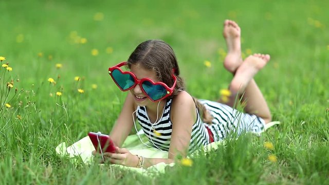Pretty girl in sunglasses with red smartphone lies on green grass. Girl in glasses lies on grass and communicates via smartphone. Female with phone. Girl with smartphone communicates through skype