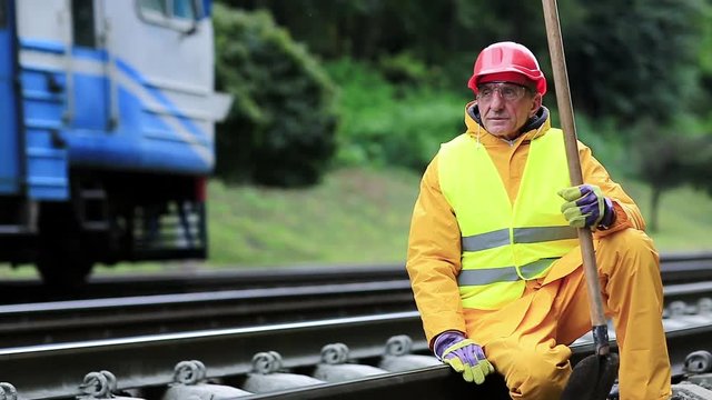 Railway worker in yellow uniform with shovel in hand sits on railway line. Railwayman in red hard hat sits on rail and looks at the train. Workman with spade on railway track. Railway construction