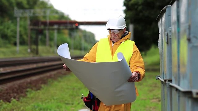 Railway worker in yellow uniform and white hard hat with wiring scheme in hands. Electrician in yellow uniform with installation diagram in hands. Railway employee near railway line