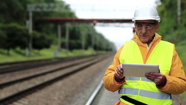 Railway worker makes notes in his tablet computer. Railway worker in yellow uniform and white hard hat with tablet pc in hands. Railway employee in yellow uniform on railway line